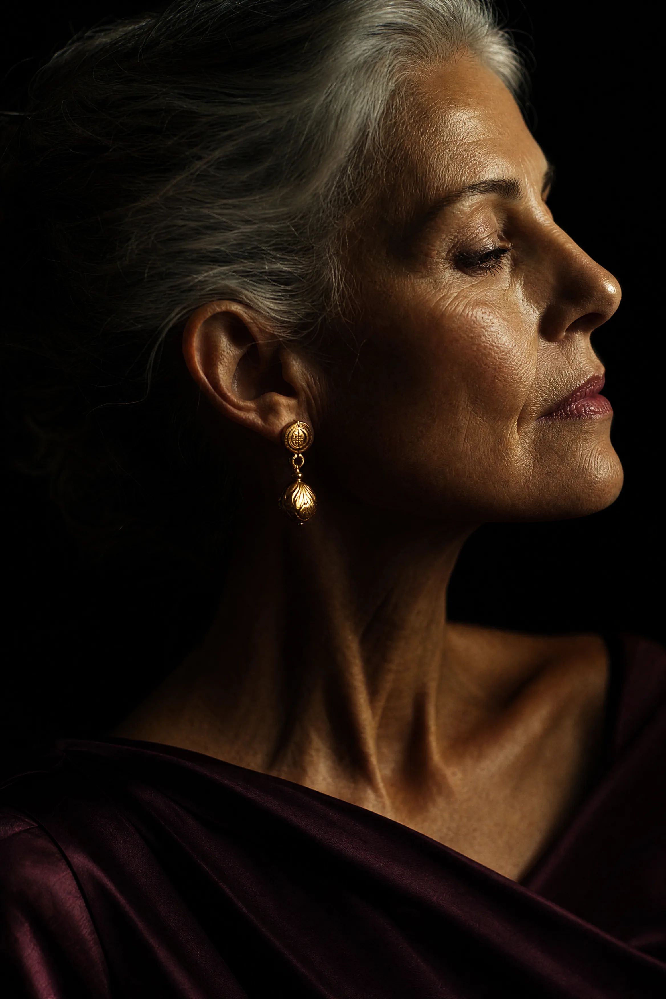 A woman with silver hair photographed in near profile against black, warm rim light tracing her brow and jaw.