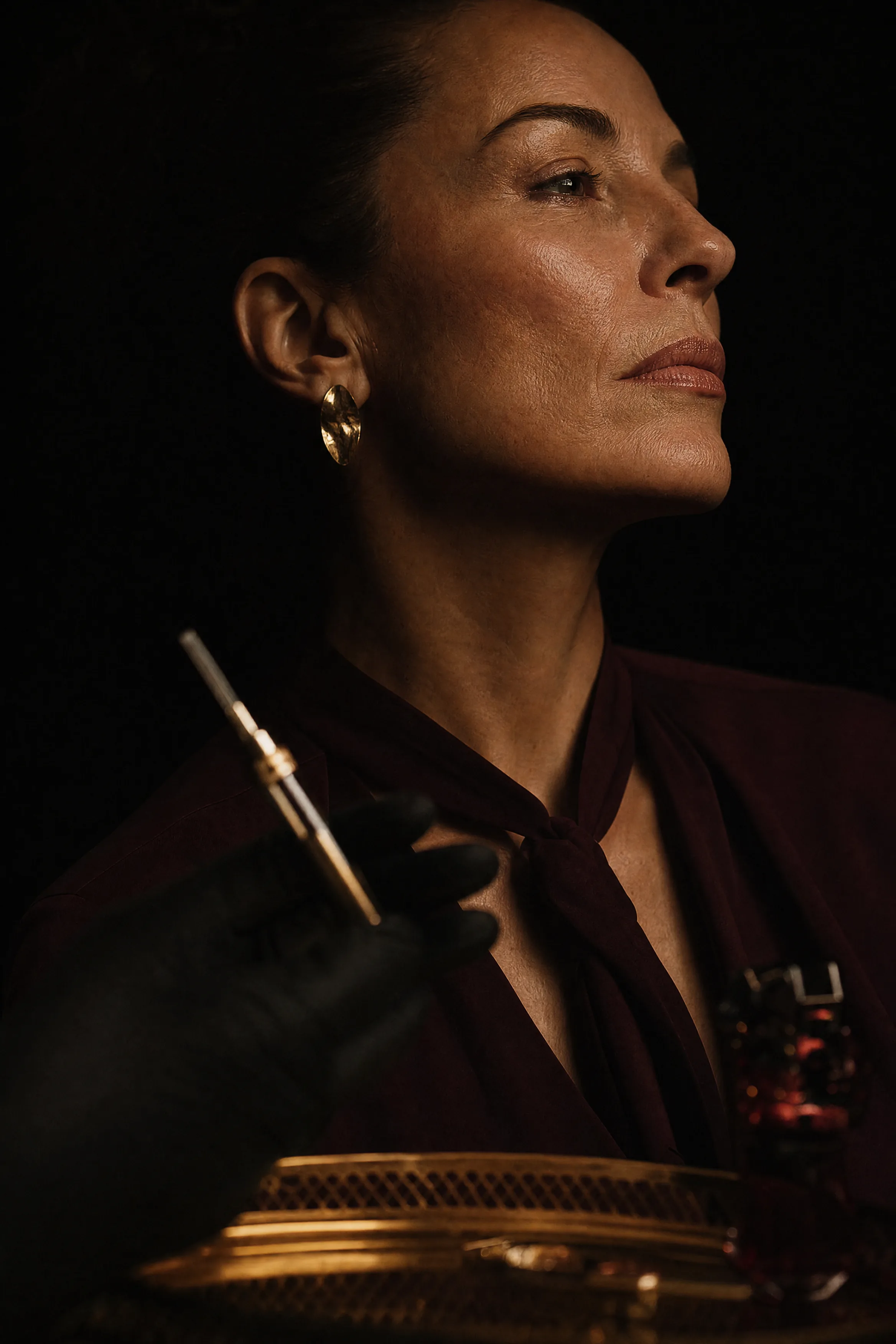 Three-quarter profile of a woman against black, jawline and lips catching a single warm highlight, a gloved hand and cannula blurred in the foreground.