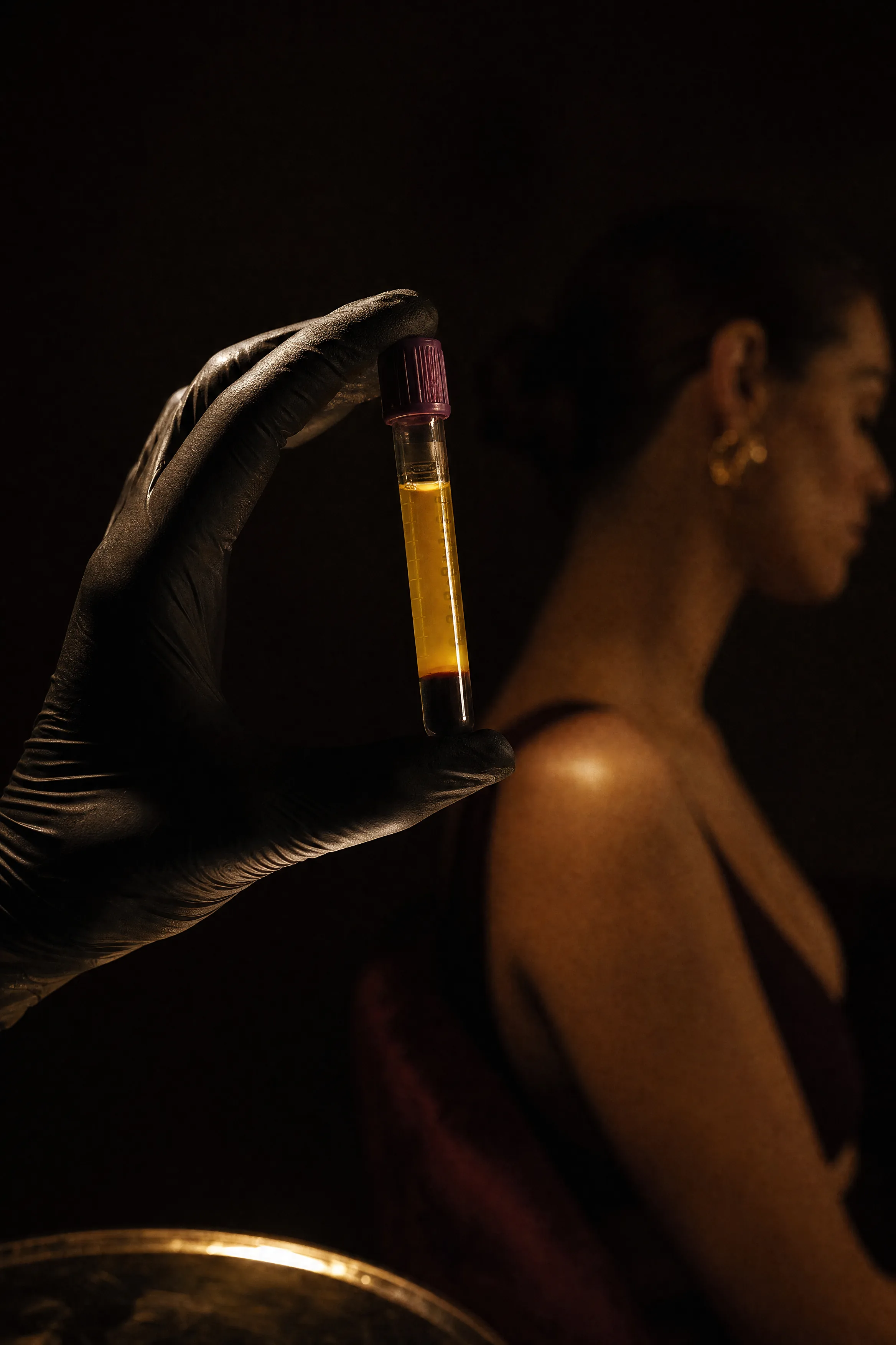 A gloved hand holds a centrifuge tube of separated golden plasma up to a single warm light against near-black.