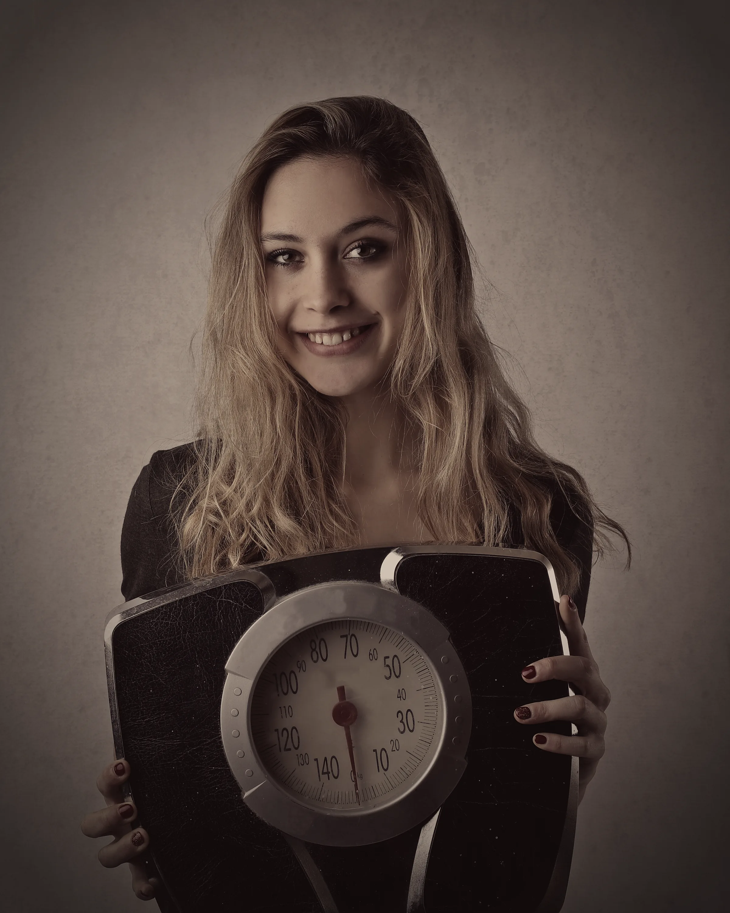 A woman stands against a plain wall holding a household bathroom scale in front of her, smiling at the camera.